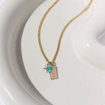 14k gold fill chain necklace featuring an initial stamped tag, three blue opal chips, and a  pearl charm, photographed on a white ceramic dish