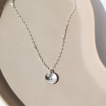 925 sterling silver Personalized Cove Necklace laid on a tan plate in the sunlight. This necklace feature our simple chain paired with the cove pendent. This pendent has the ability of being stamped with any initial of your choice.