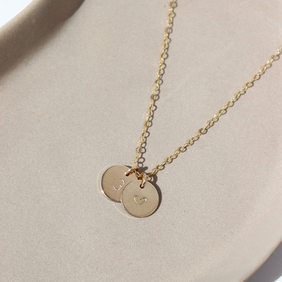 14k gold fill Tiny Monogram Disc Necklace laid on a gray plate in the sunlight. This necklace features our simple chain with a disc pendent with a heart stamped on it as well as an inital.