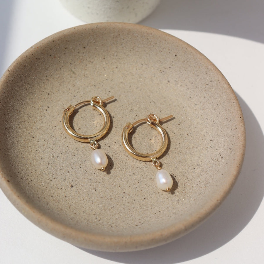 14k gold fill classic pearl hoops laid on a tan plate in the sunlight.