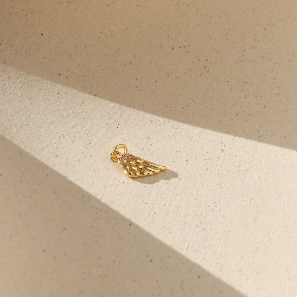 Gold angel wing charm with sparkling crystal details, photographed on a warm neutral surface with soft natural light and shadows.