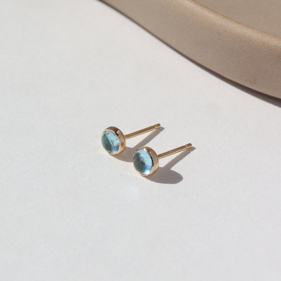 4mm Blue Topaz gemstones set in a 14k gold fill bezel stud earring and placed on a white plate for display. Sold as a pair and perfect for single or double pierced ears.