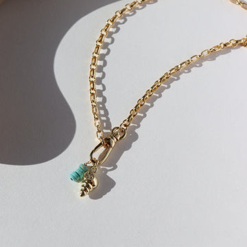 14k gold fill anklet with a swivel push-gate clasp holding a seashell charm and a turquoise charm, photographed on a sunny table