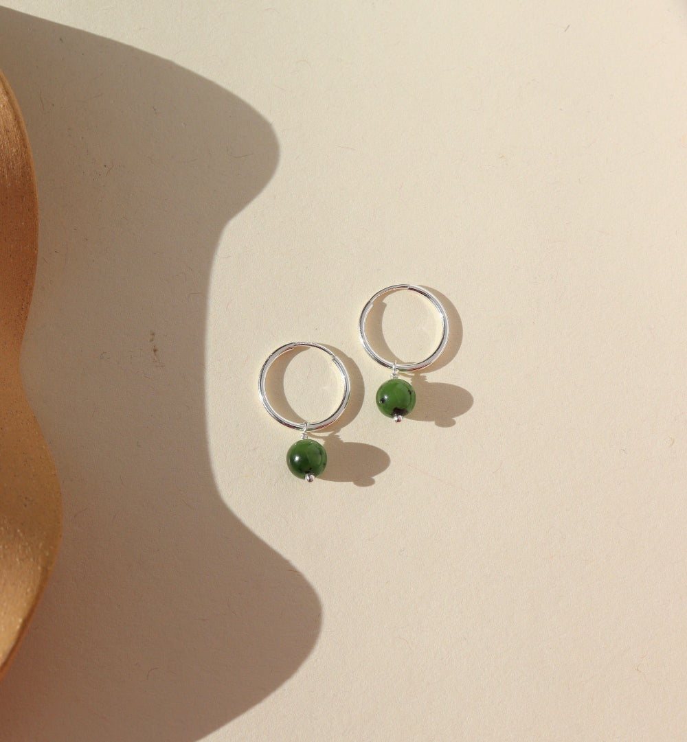 A sterling silver 14mm hoop with a Jade ball gemstone dangling from it. Earrings are displayed on a cream background. 