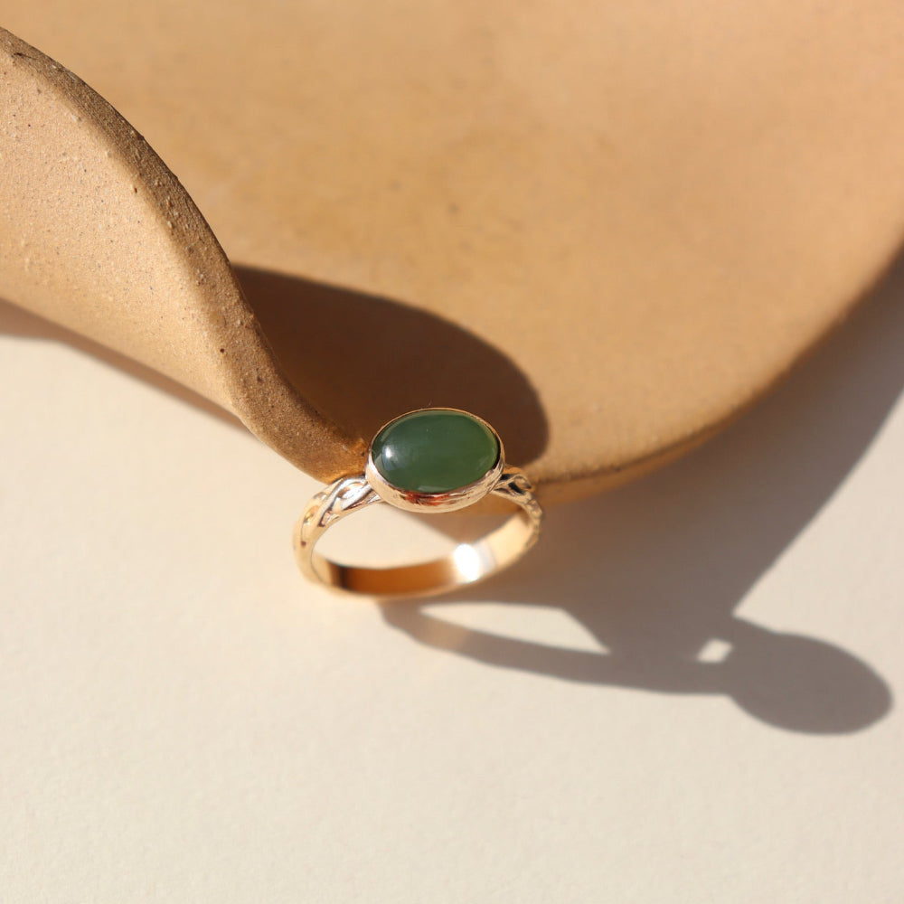 A 14k gold fill entwined band with a 8x10 jade gemstone in a gold bezel. The ring is displayed on a orange dish. 