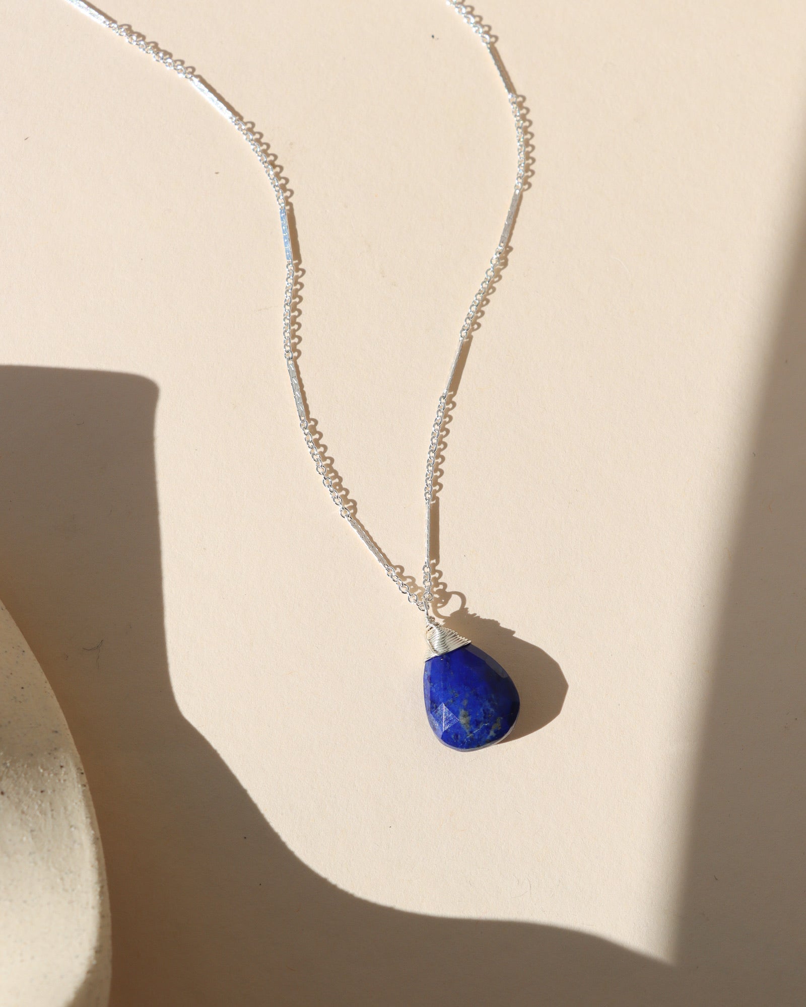 A silver chain necklace with a wire-wrapped, teardrop-shaped lapis lazuli pendant lying on a beige background.