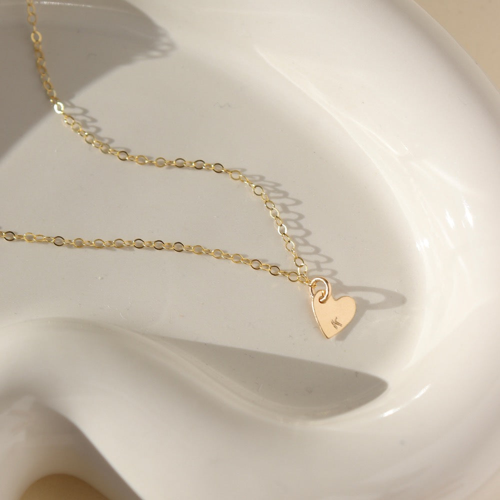 a dainty heart charm with an initial stamped on them, on a necklace chain, photographed on a ceramic dish