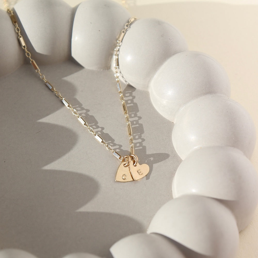 two dainty heart charms with initials stamped on them, on a necklace chain, photographed on a ceramic dish