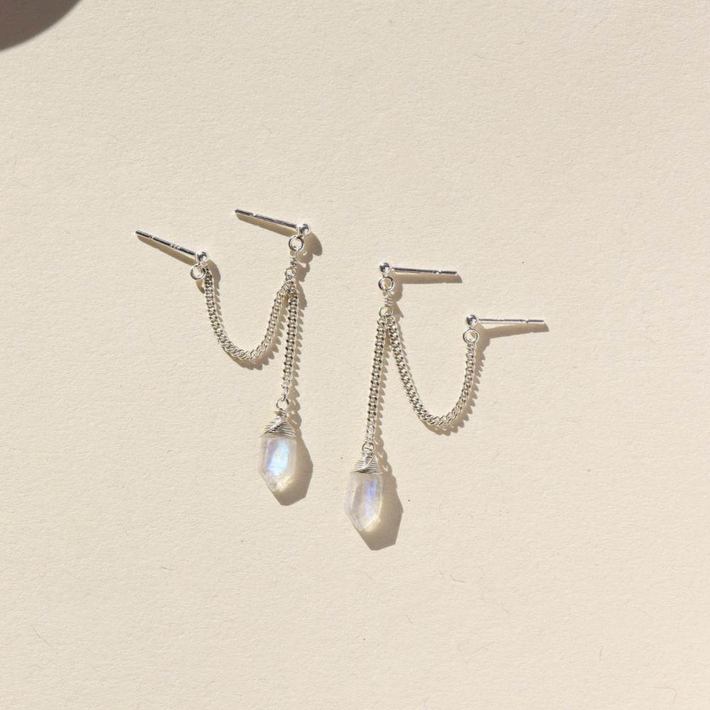 Two small studs with silver curb chain and a hexagon moonstone attached to the end of each earring. Earrings are displayed on a cream backdrop. 