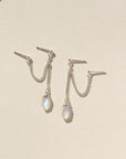 Two small studs with silver curb chain and a hexagon moonstone attached to the end of each earring. Earrings are displayed on a cream backdrop. 