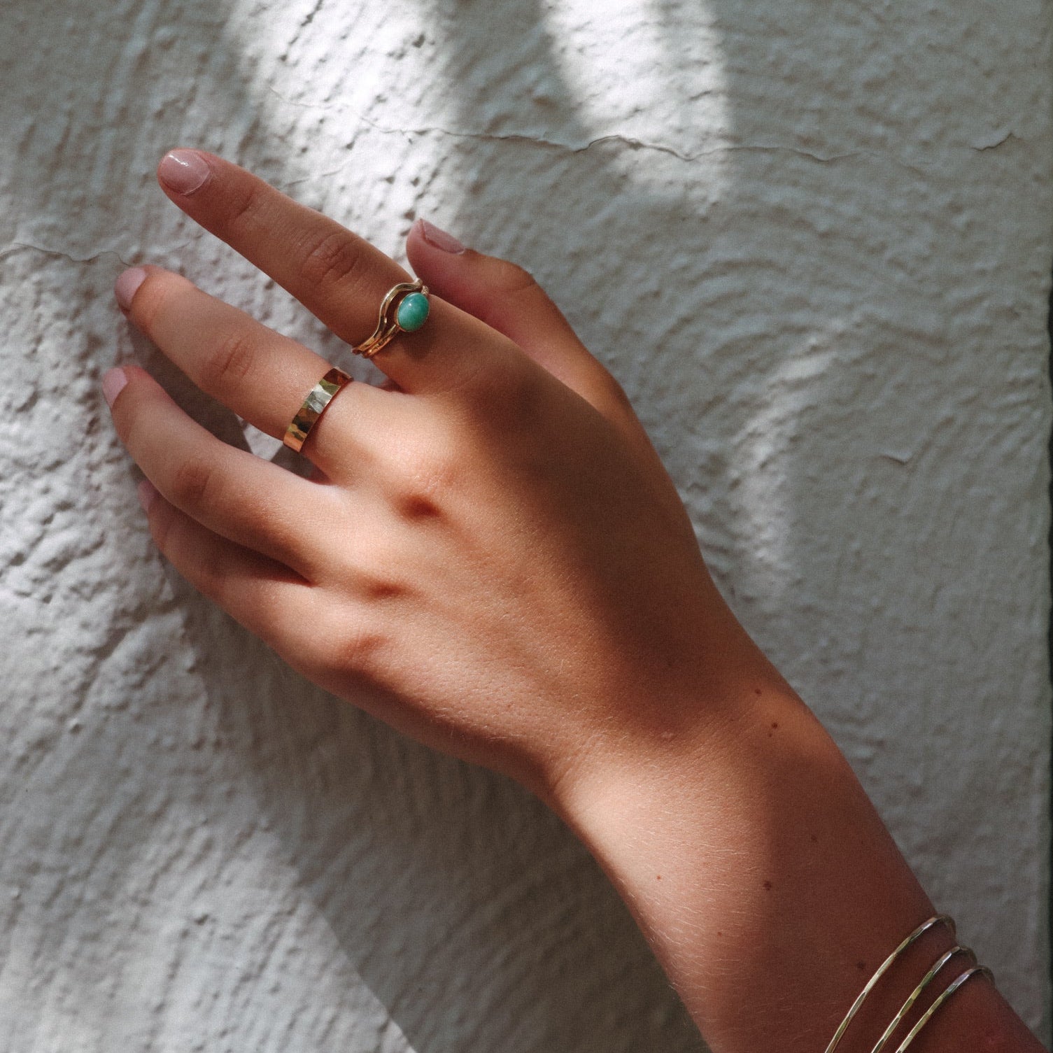 Hand wearing a 14k gold fill turquoise statement ring with a polished gold band and slim stacking bangles, photographed against a textured white wall in natural sunlight.
