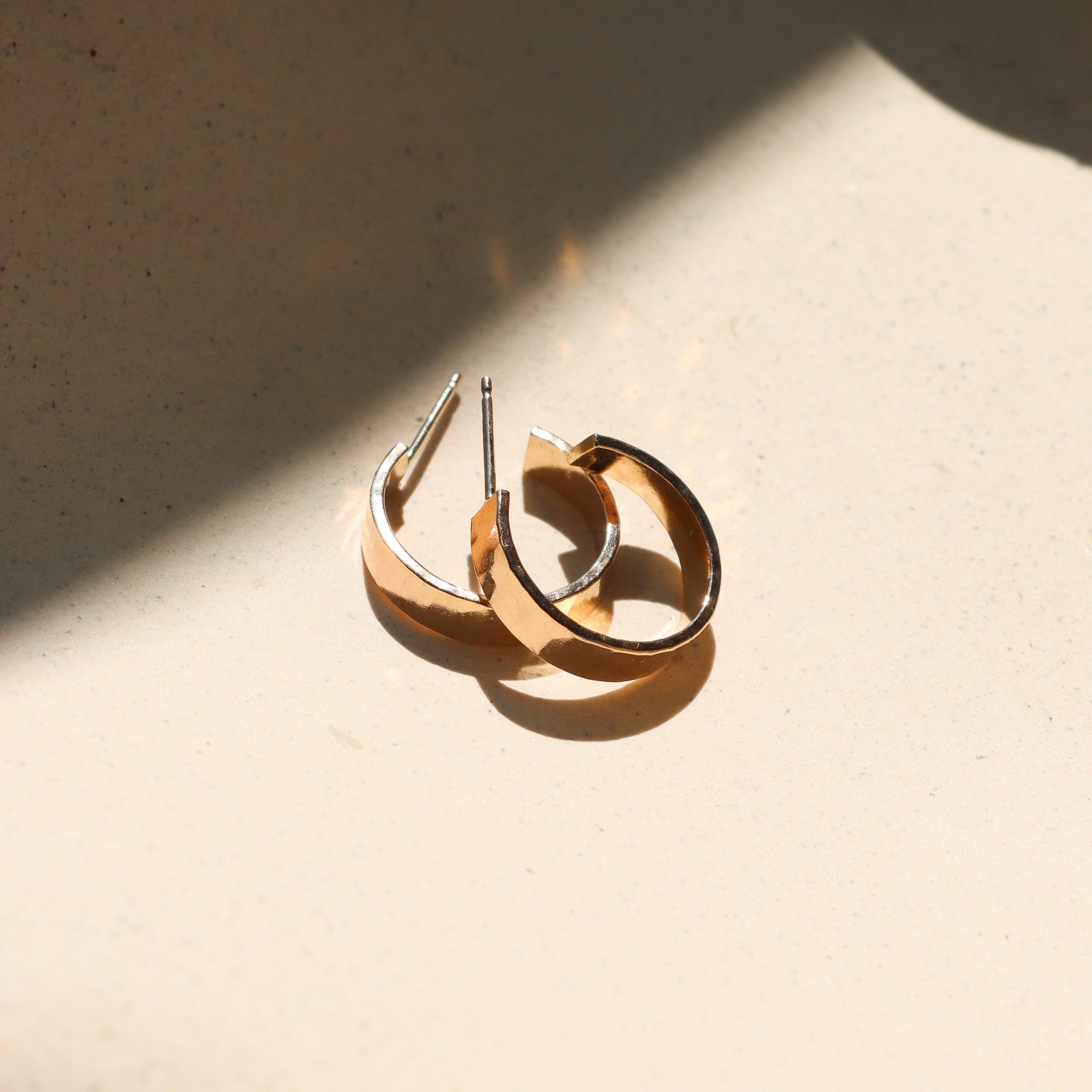 A pair of thick, polished gold hoop earrings resting on a textured beige surface under dramatic, direct sunlight with soft shadows.