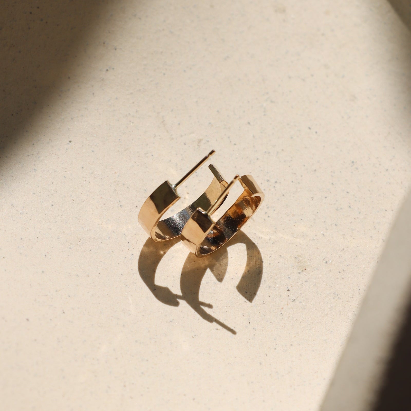 A pair of gold-toned geometric hoop earrings resting on a textured beige surface with dramatic sunlight and shadows.