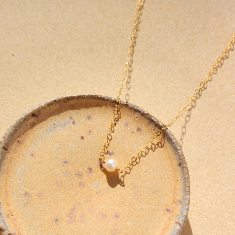 14k gold fill Mini Pearl Necklace laid on a tan bowl in the sunlight. This Necklace features a mini pearl with connected by the simple chain.