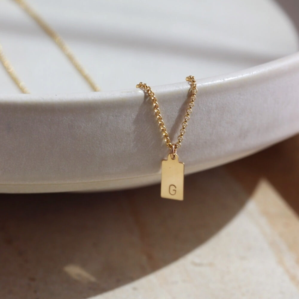 A gold rolo chain with a stamped mini tag with the letter "G". Necklace is displayed over a stone dish. 