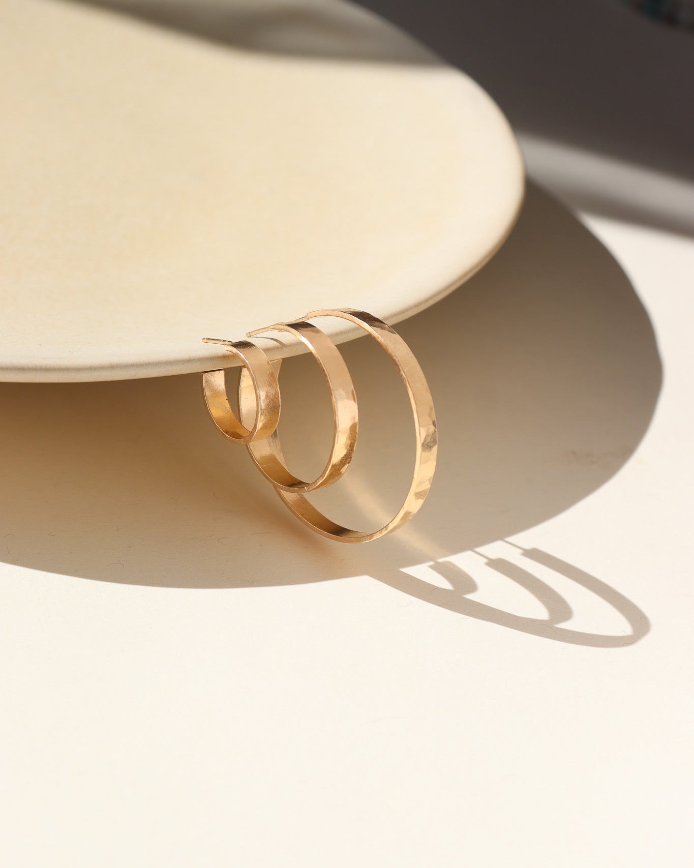 A thick hammered hoop offered in three sizes (small, medium, and large) displayed hanging on a tan dish. 