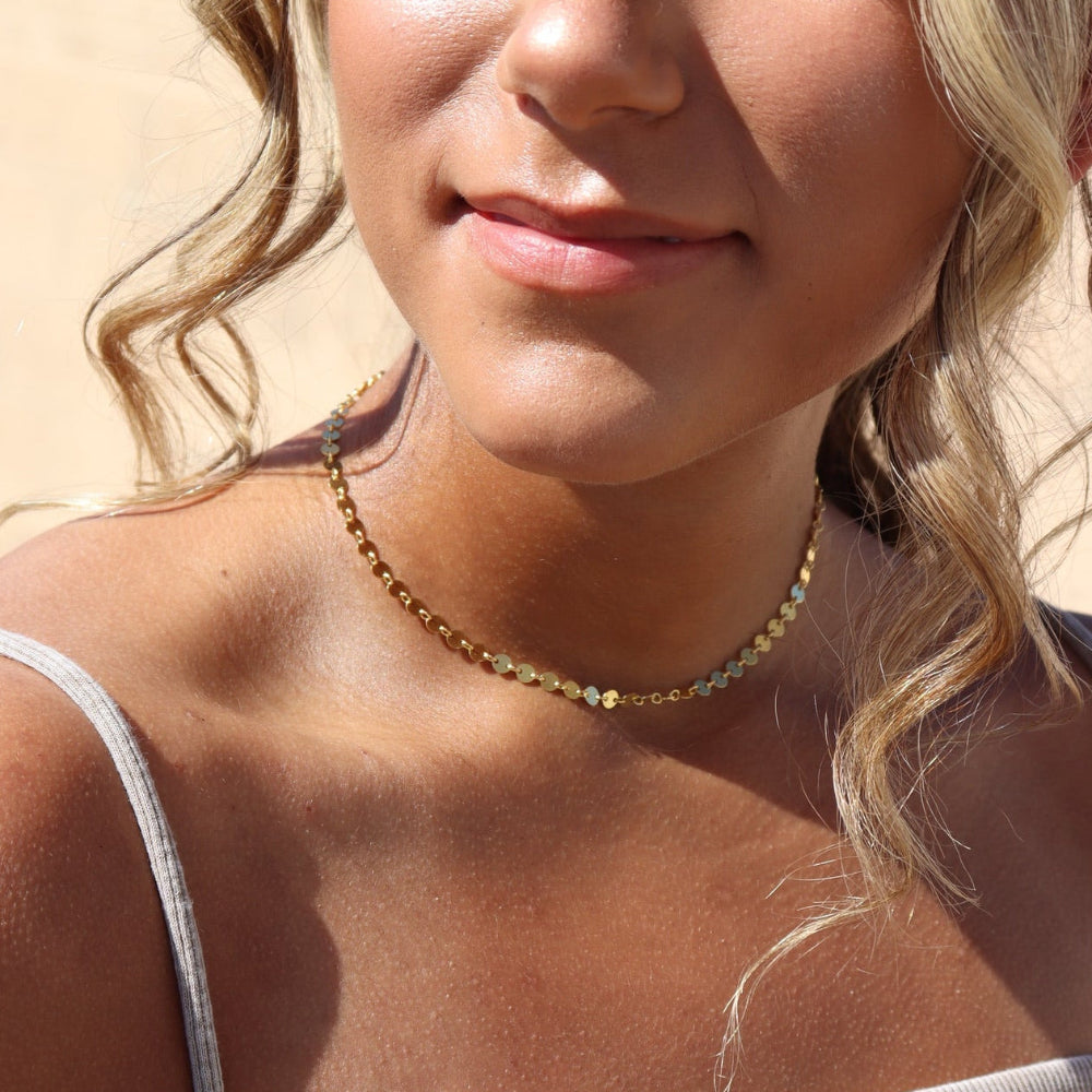 Woman with blonde hair and a necklace, wearing a beige top, outdoors.