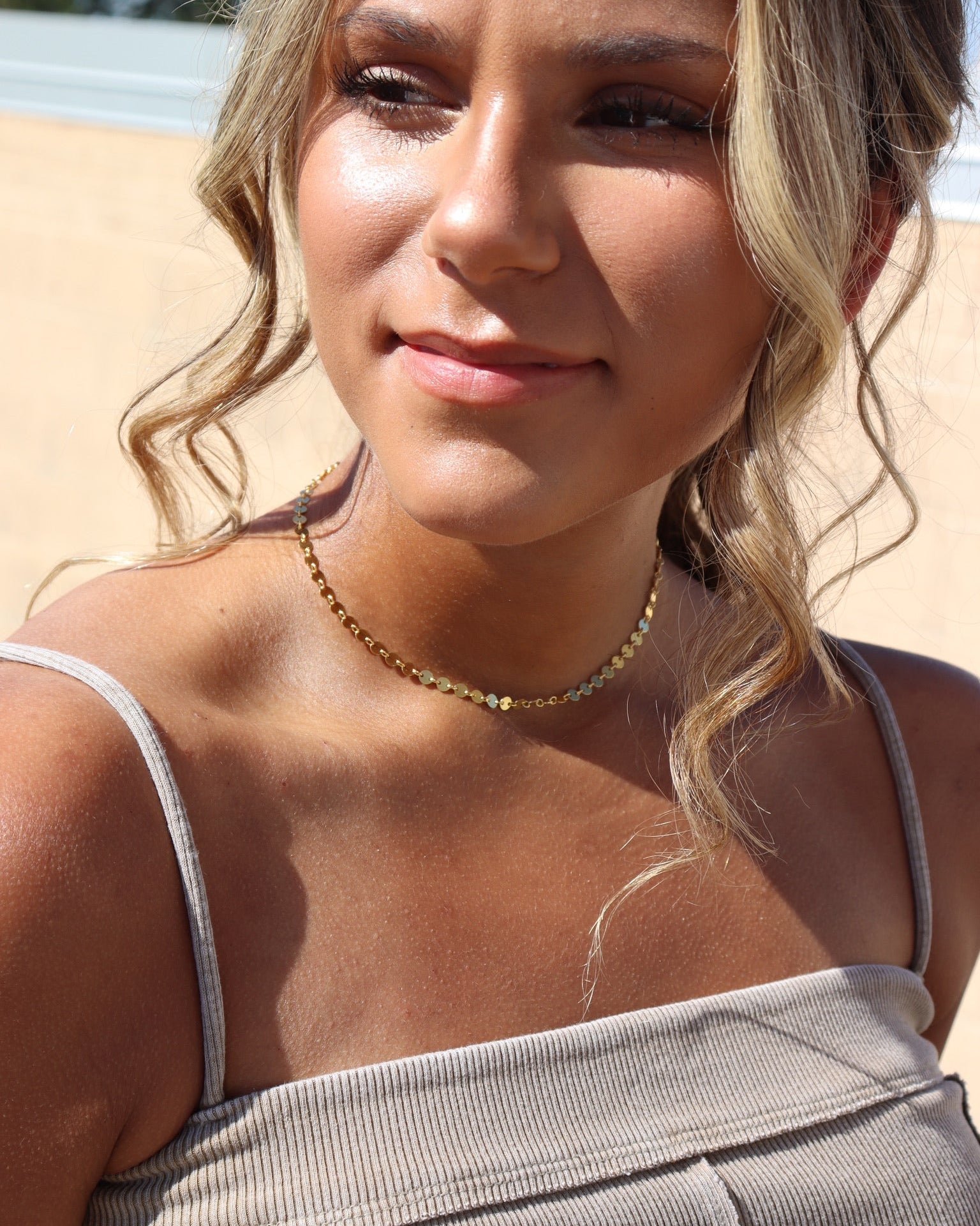 Woman with blonde hair and a necklace, wearing a beige top, outdoors.