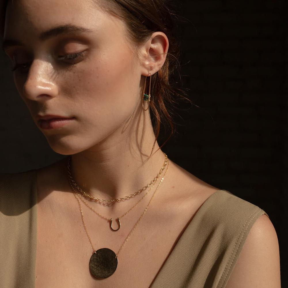 Brunette model wearing a 14k gold fill Lucky Charm Necklace. This Necklace features a horseshoe shape with hammering on one side. The horseshoe meaning lucky is connected with the simple chain.