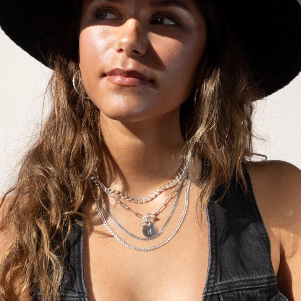 Sterling silver chain link necklace with a toggle clasp and monogrammed "B" flat disk charm, photographed on a model with long brown hair, wearing silver jewelry, a black denim vest, and a black cowboy hat