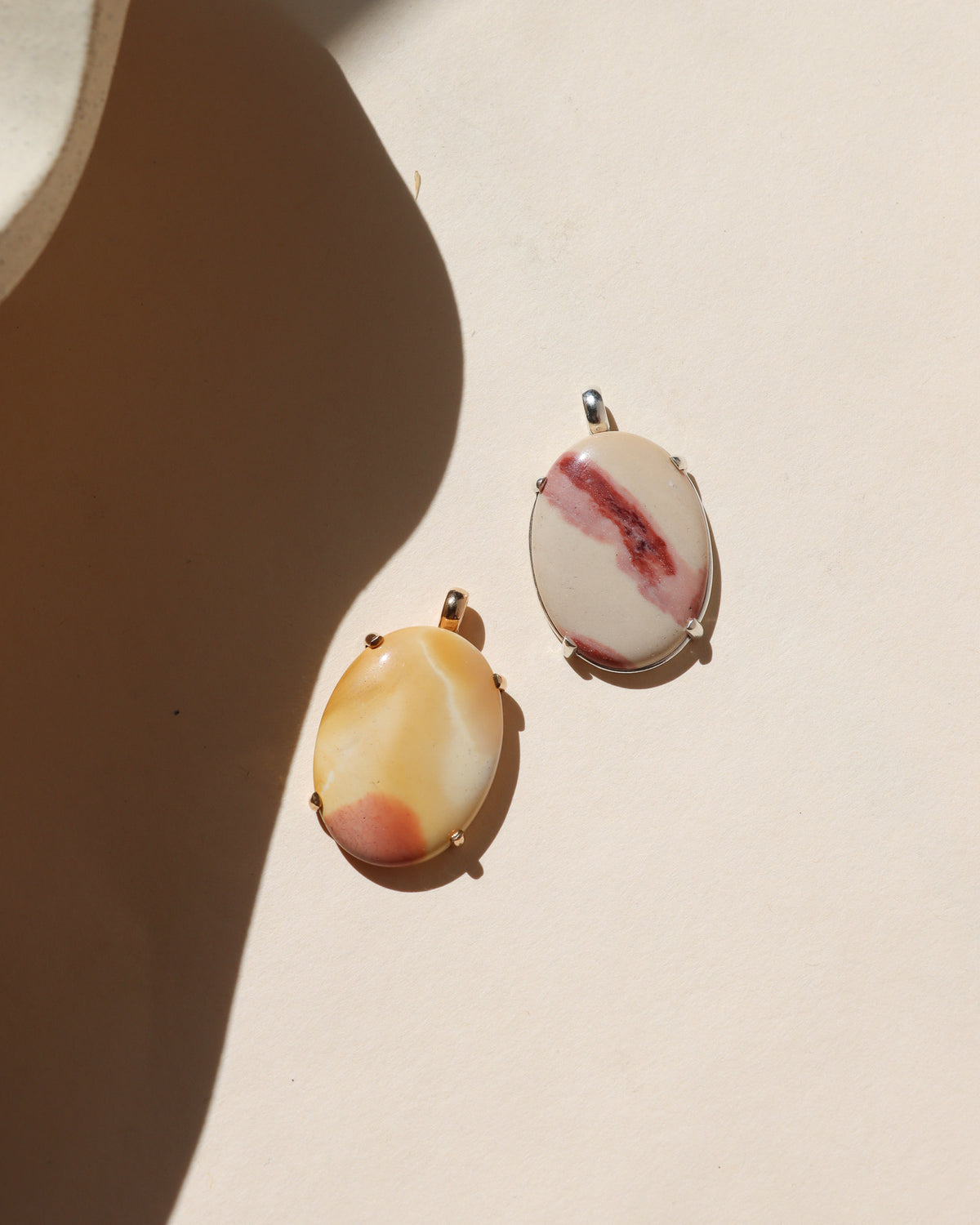Two oval stone pendants in warm cream, yellow, pink, and red tones set in 14k gold fill and sterling silver, photographed on a cream background in warm sunlight.