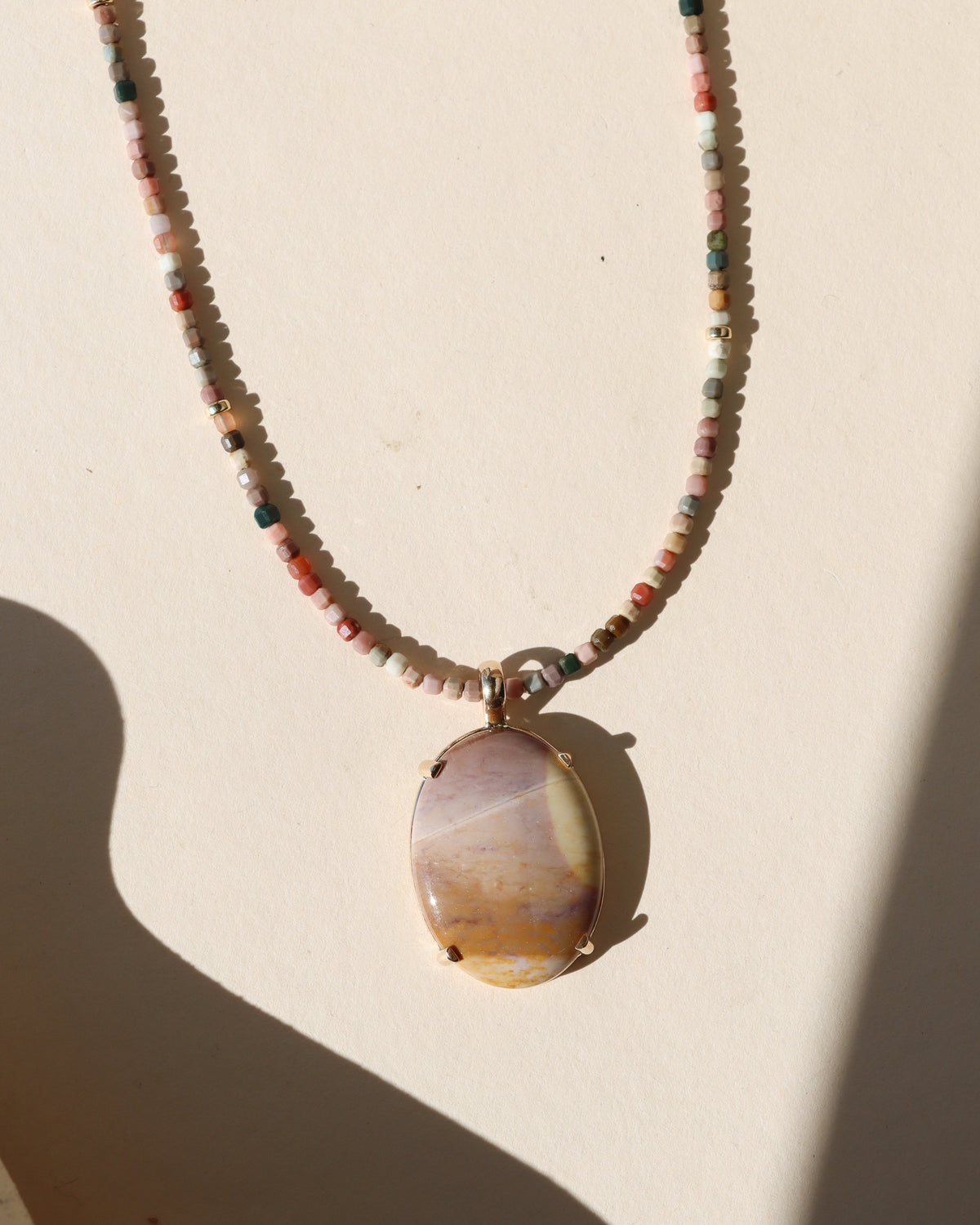 Multicolored beaded necklace featuring an oval cream, lavender, and golden stone pendant in a gold setting, photographed in warm sunlight.
