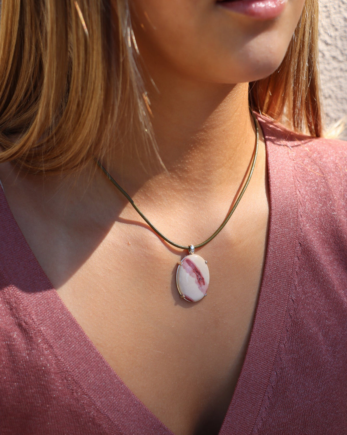 Oval cream and red stone pendant on an olive cord necklace, worn with a plum top in warm sunlight.
