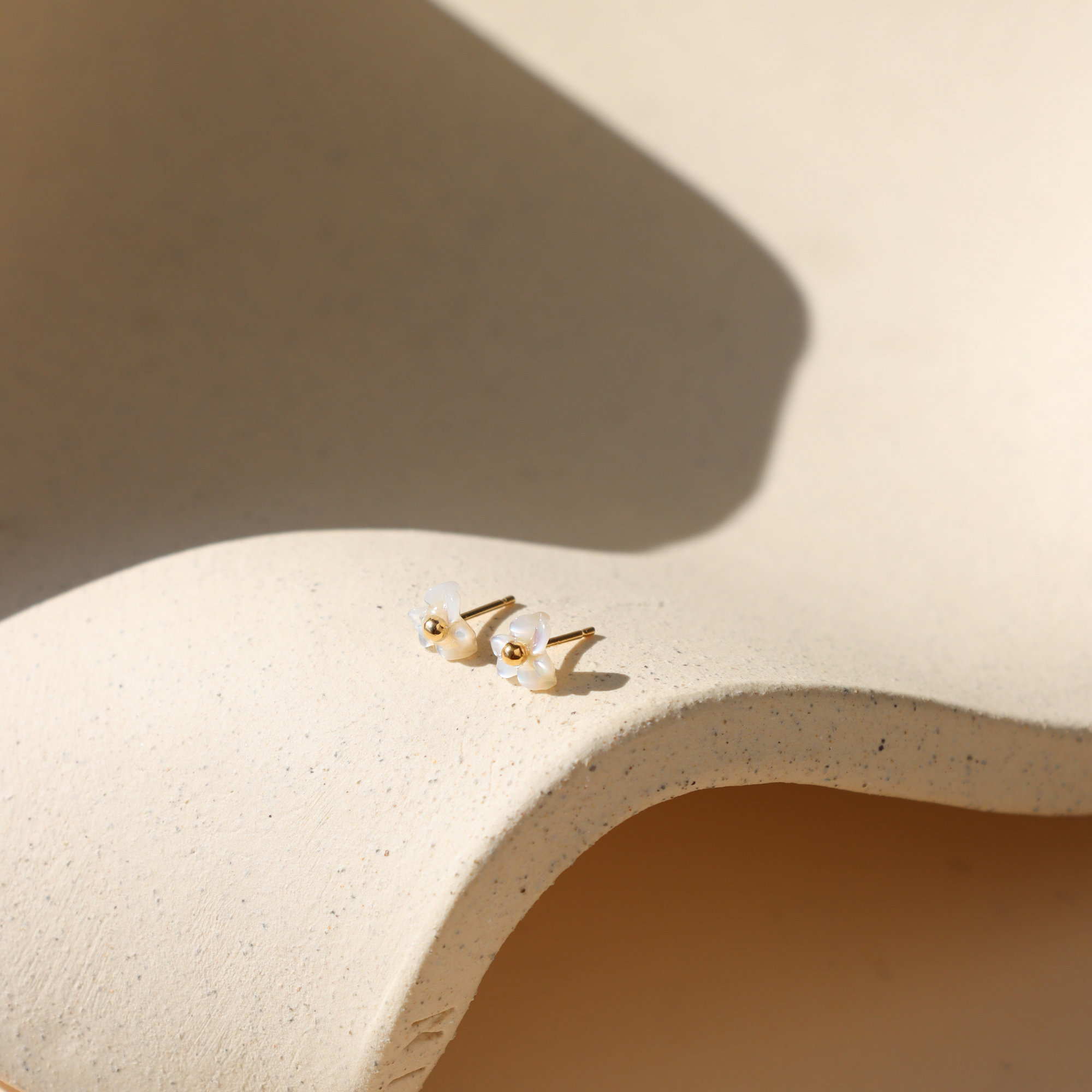 Pair of mother-of-pearl flower stud earrings with polished 14k gold fill centers resting on a textured cream ceramic display in warm natural light.