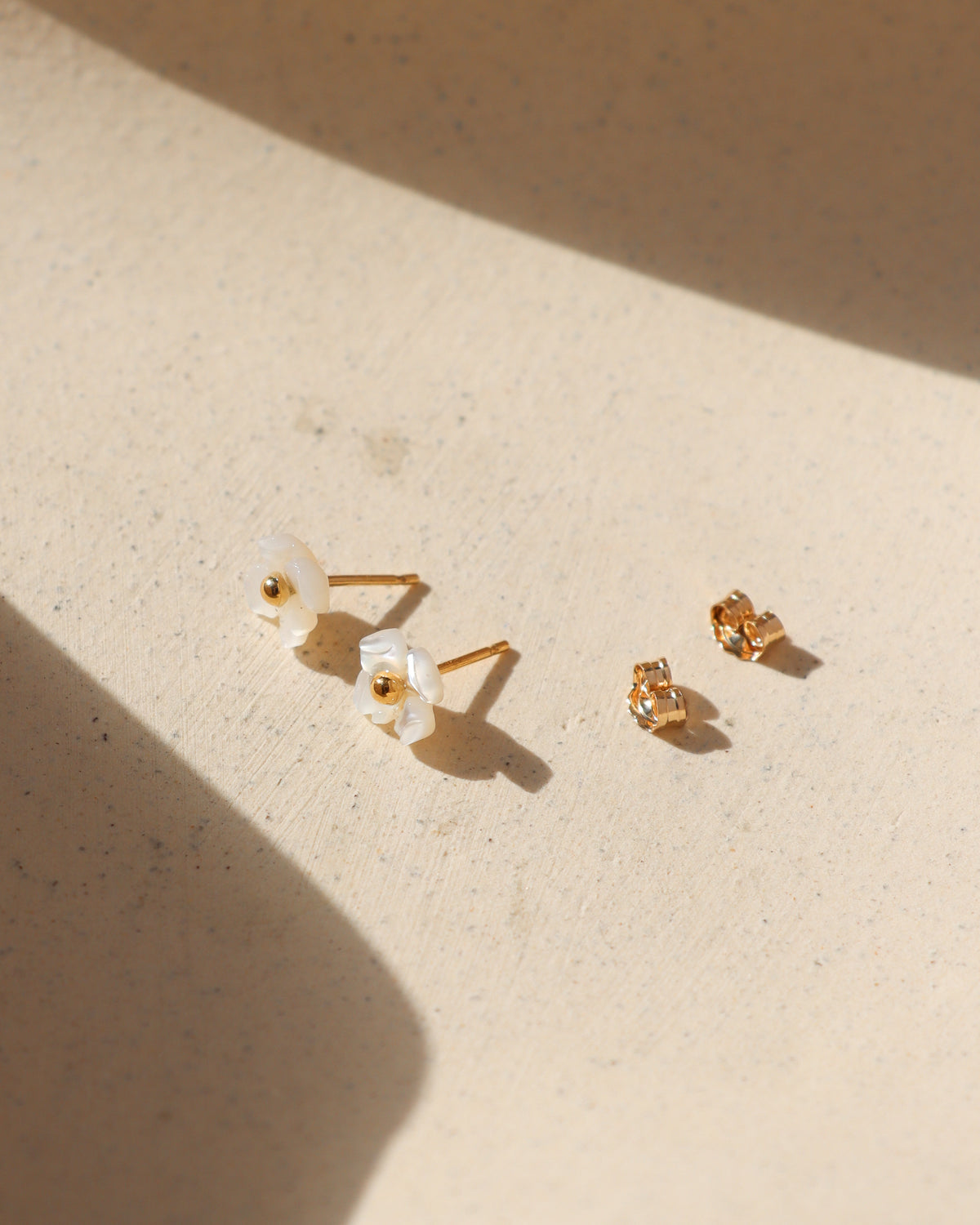Pair of gold stud earrings featuring delicate white floral petals and gold centers, arranged on a sunlit cream stone surface with soft shadows.