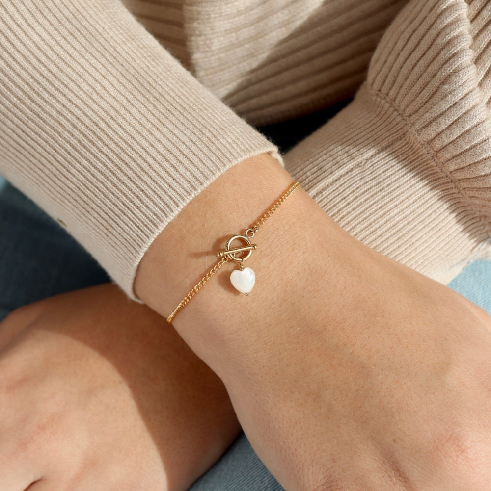 Model wearing the mother of pearl toggle bracelet. This bracelet features a gold curb chain with a toggle clasp opening with a mother of pearl heart at the end. 