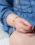 a little girl wearing 14k gold fill jewelry by Token Jewelry, including a spiral ring with a heart charm and the Starlight Bracelet. She's wearing a blue denim dress and sitting on a white couch