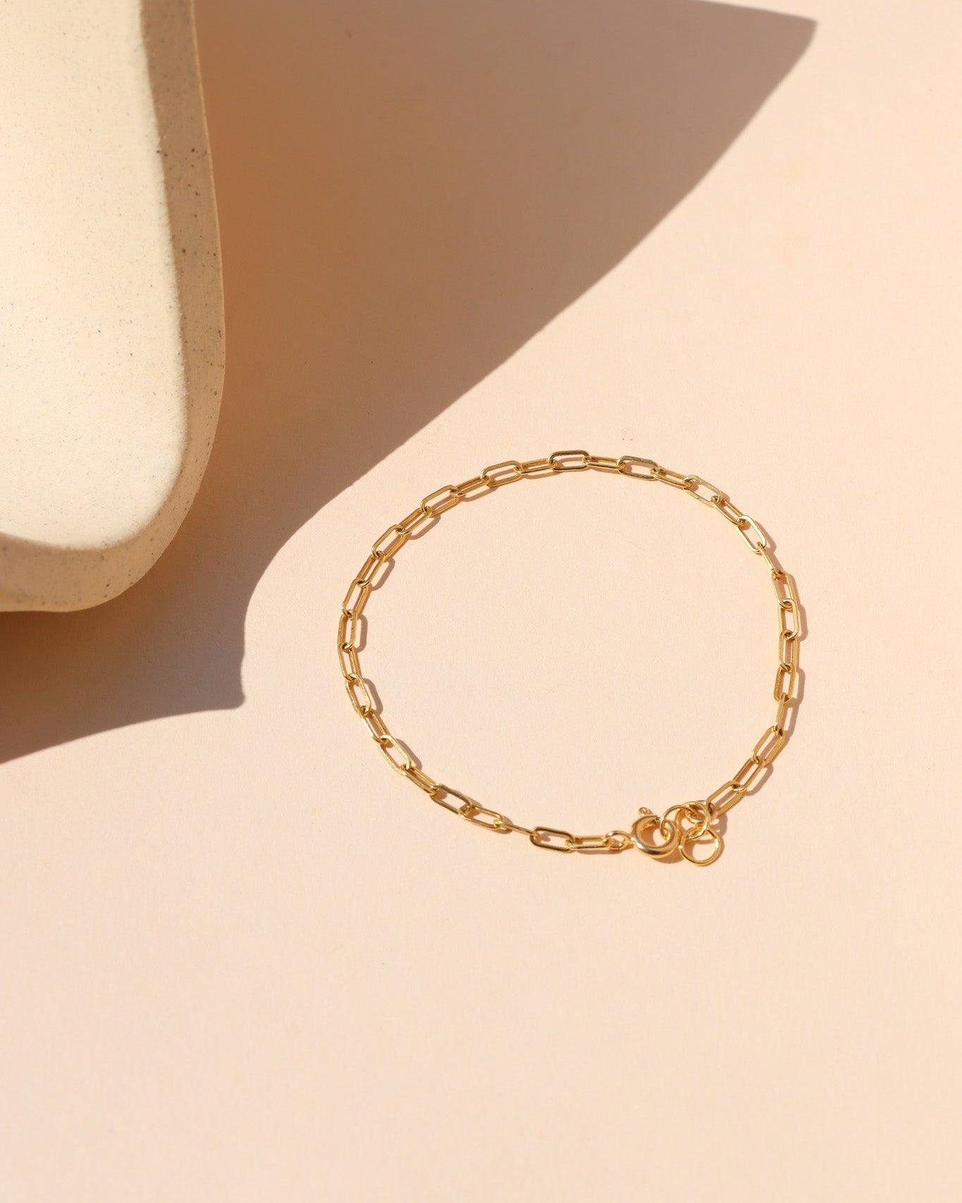 A delicate gold paperclip chain bracelet displayed on a warm beige surface with a geometric shadow.