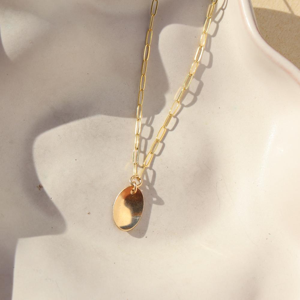 14k gold fill Olivia Necklace laid on a tan plate in the sunlight. This necklace features out cosette chain that looks like paper clips linked together as well as an oral charm that is hammered on one side