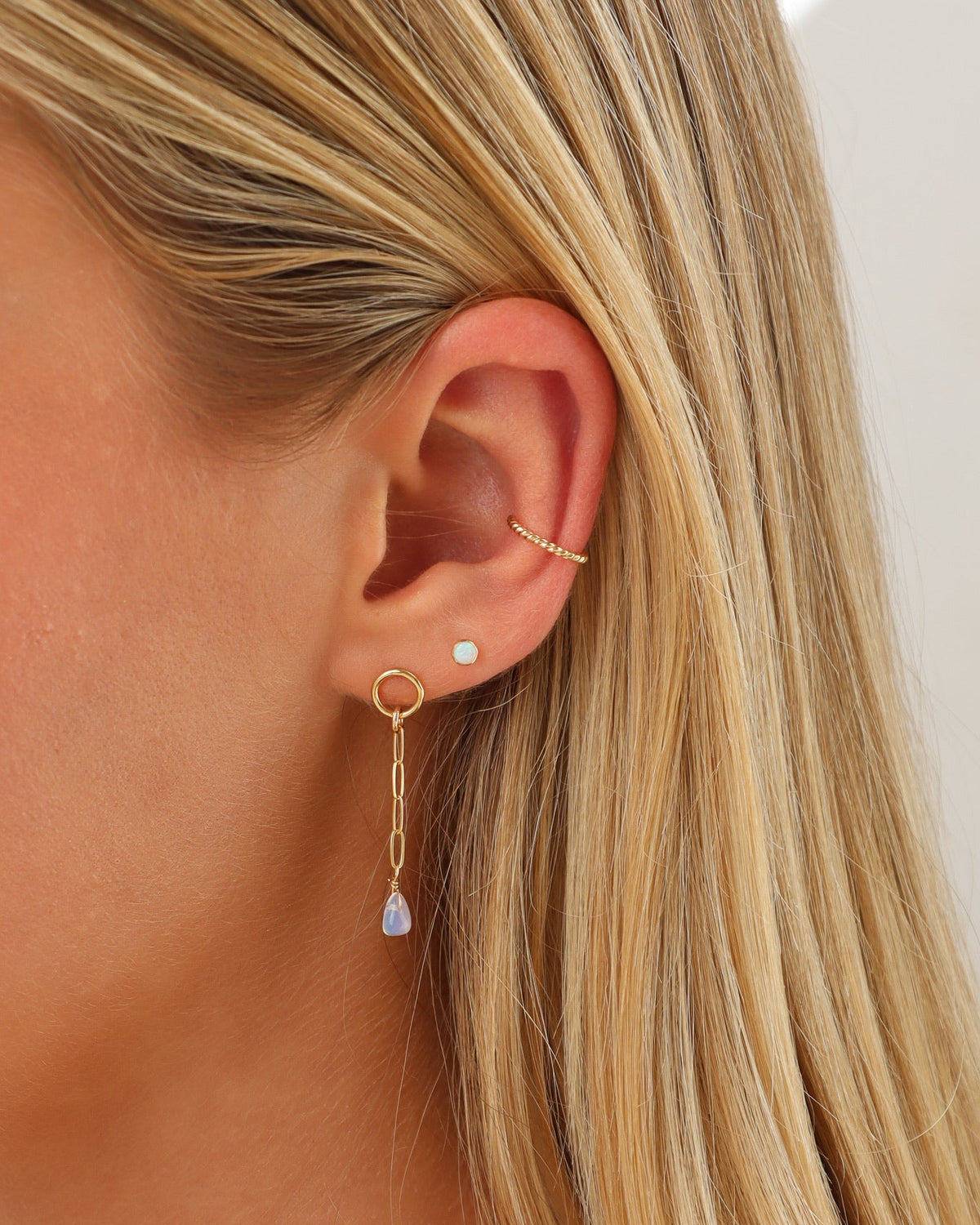Close-up of a woman wearing a twisted gold spiral ear cuff, a petite opal stud, and a delicate gold chain drop earring finished with a teardrop opal gemstone.