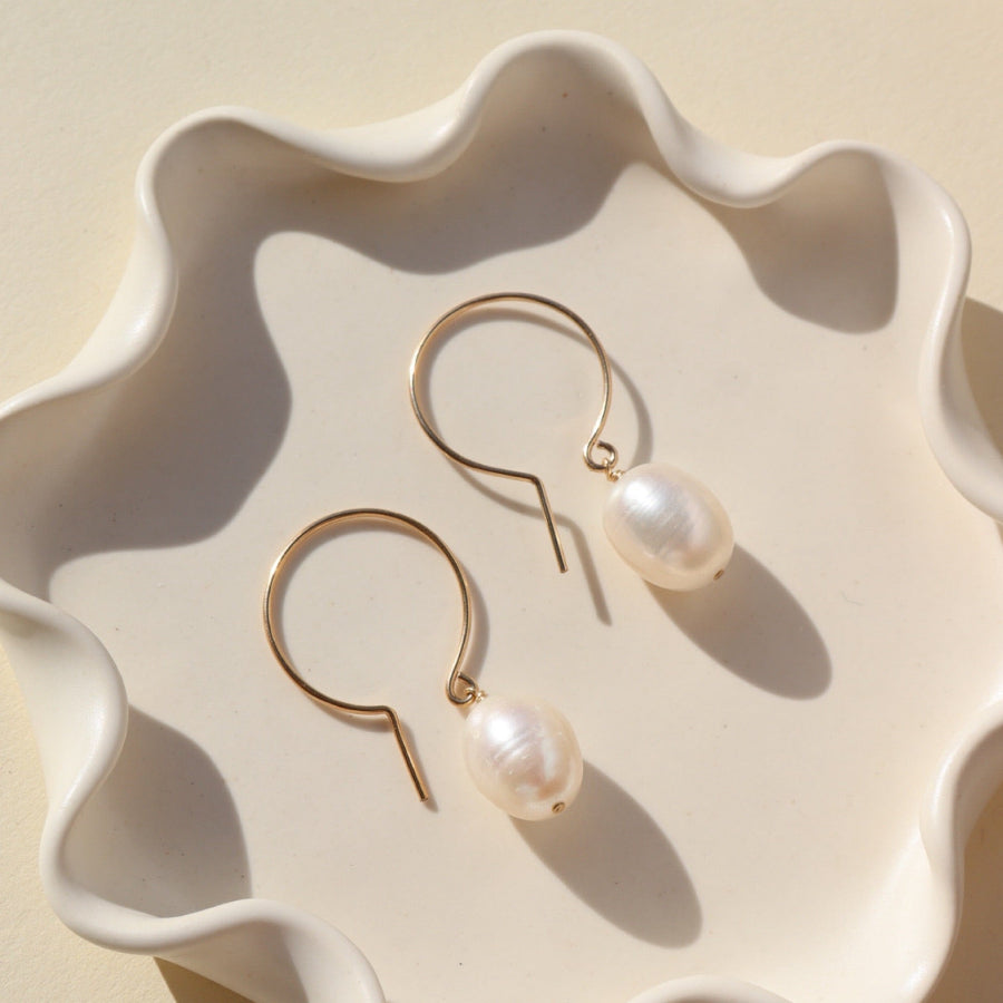 A pair of pearl earrings resting on a white, wavy ceramic dish in natural light.