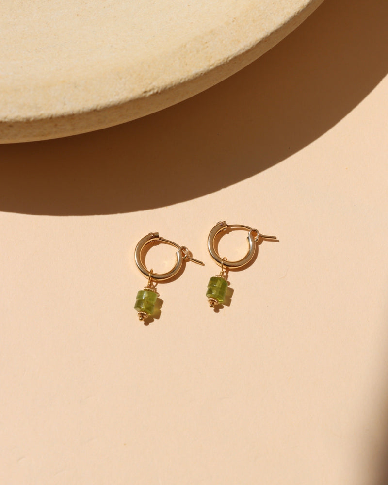 Petite gold hoop earrings with natural green peridot gemstone drops, photographed in warm sunlight.