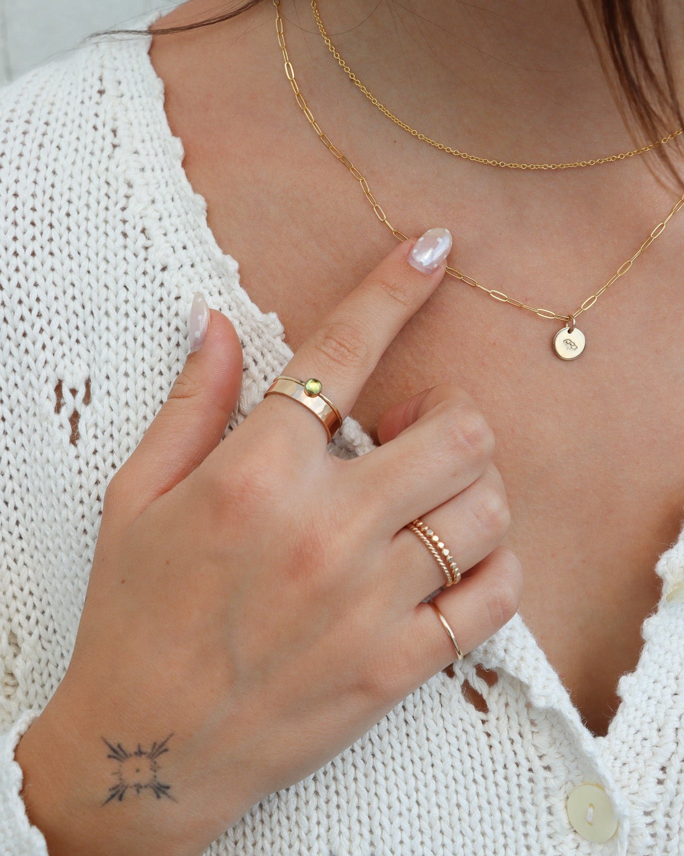 A close-up lifestyle shot of a woman wearing delicate gold jewelry, including layered necklaces and multiple rings, paired with a white knit top.