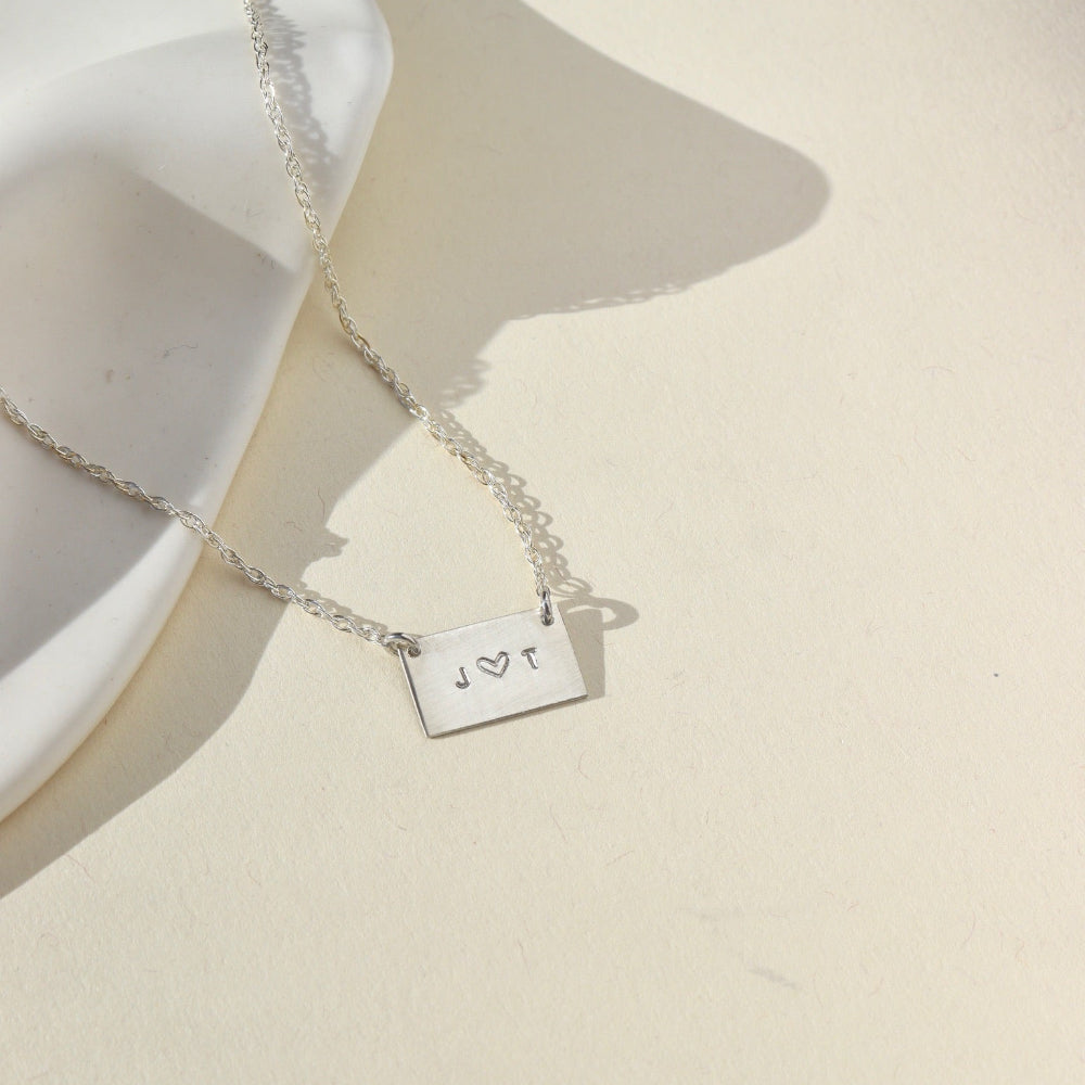 Sterling Silver necklace with a thin rolo chain with a silver smooth rectangle pendant with a "J" and a "T" stamped in the middle of it. The necklace is displayed on a cream backdrop. 