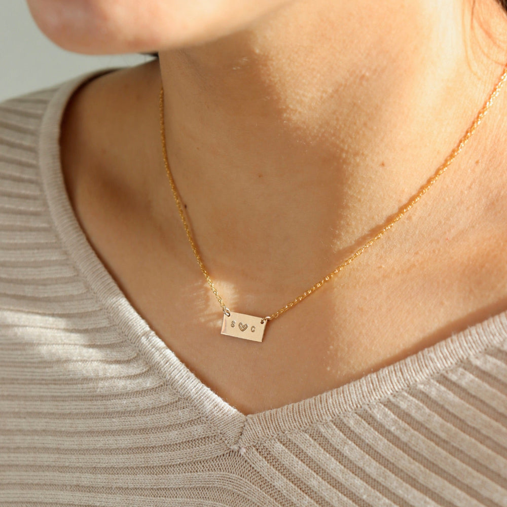 Model wearing 14k gold fill necklace with a thin rolo chain with a gold smooth rectangle pendant with a "S" and a "C" stamped in the middle of it. 