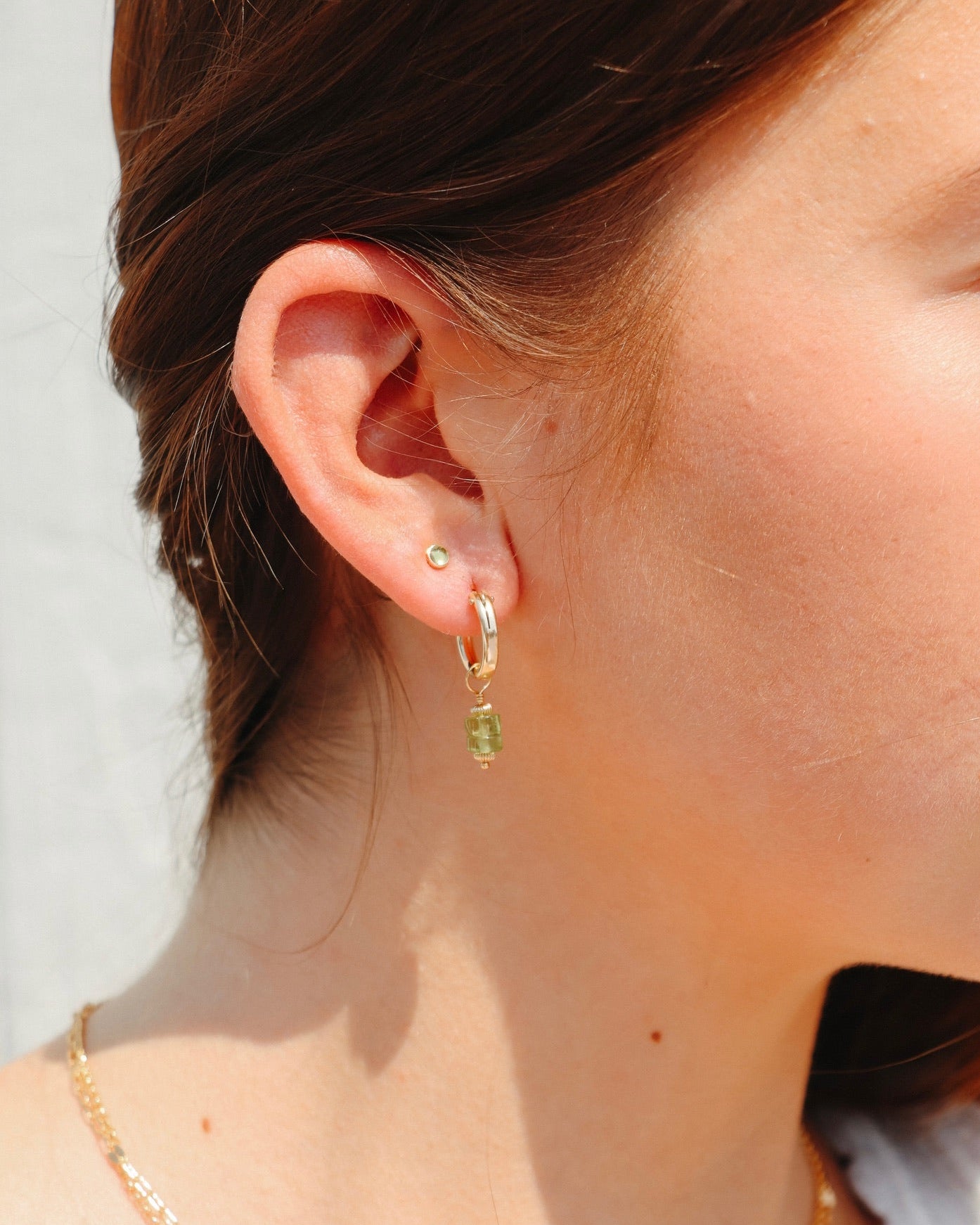 Model wearing a petite peridot birthstone stud earring in a gold setting, paired with a green peridot drop hoop earring.