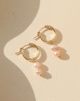 Small gold hoops featuring two pink pearls dangling from them. Earrings are displayed on a cream backdrop.