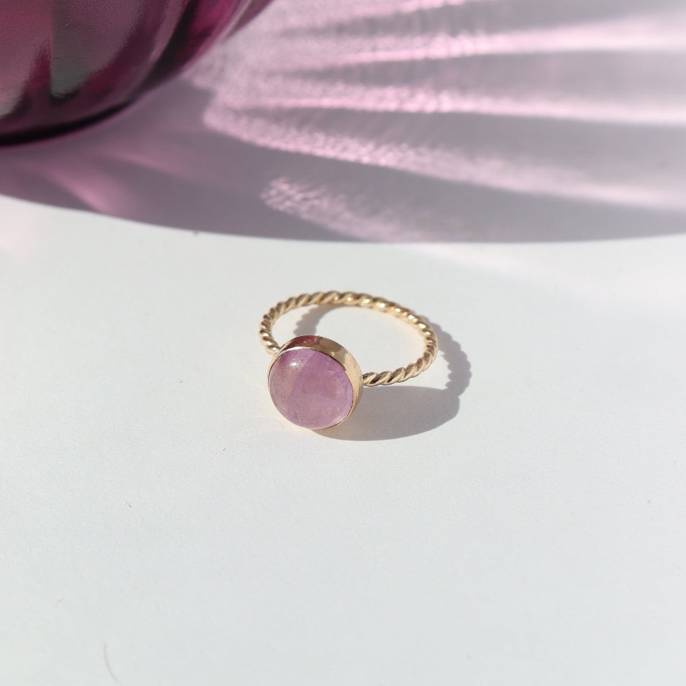 14k gold filled Pippa Ring laid on a white paper in the sunlight. This ring features a sprial band with a pink kunzite gemstone.