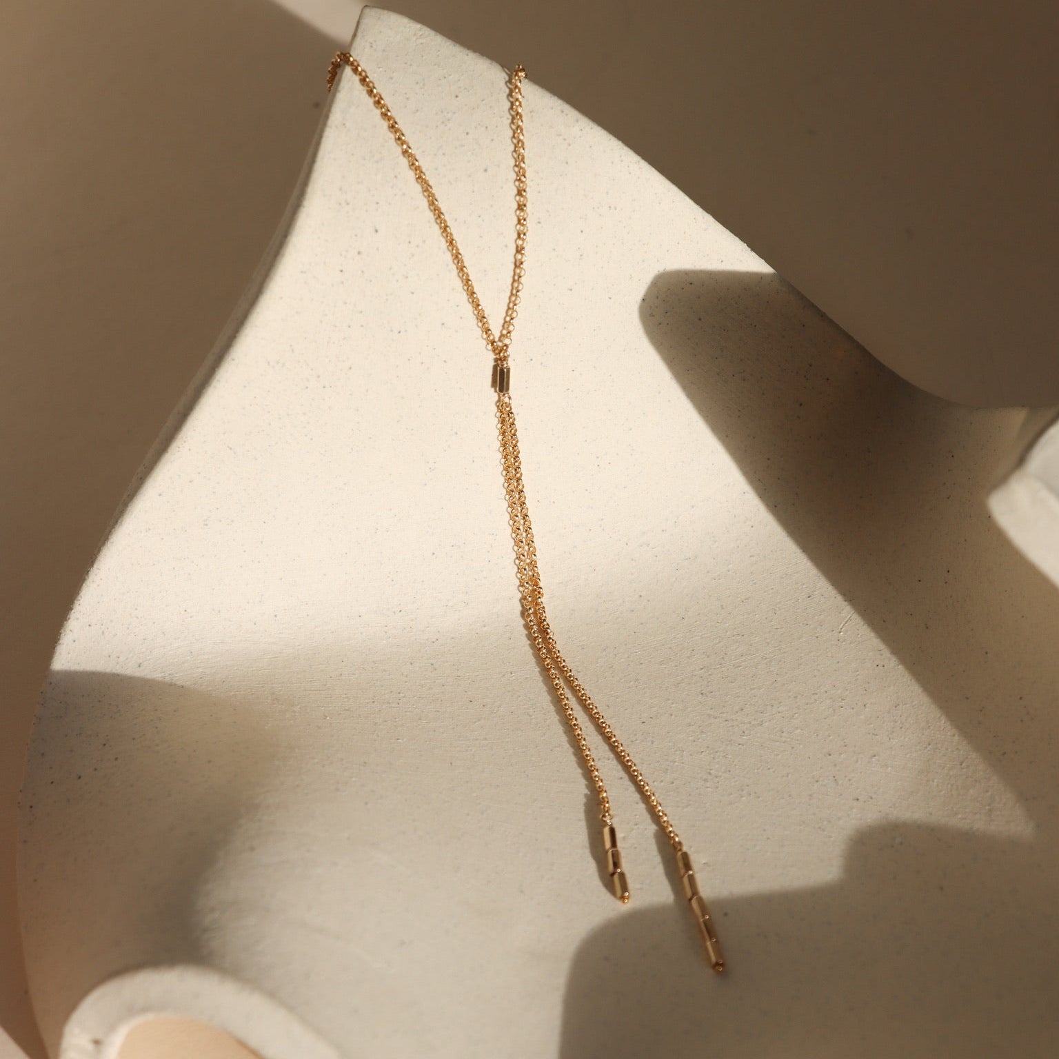 Remy chain lariat necklace in gold with a fine chain and elongated drop accents, styled on a sculptural neutral backdrop.