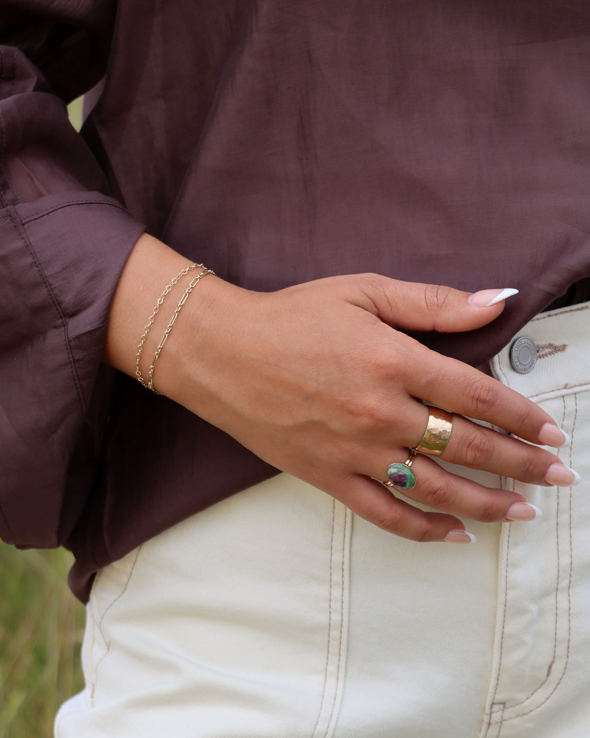 Model wearing an oval green and pink gemstone ring stacked with a wide hammered gold ring and delicate gold chain bracelets.