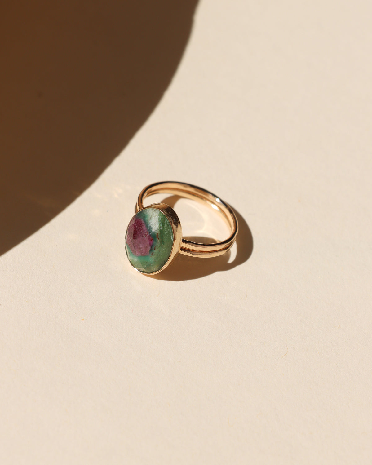 Gold ring with an oval green and deep pink gemstone photographed on a cream background in warm sunlight.
