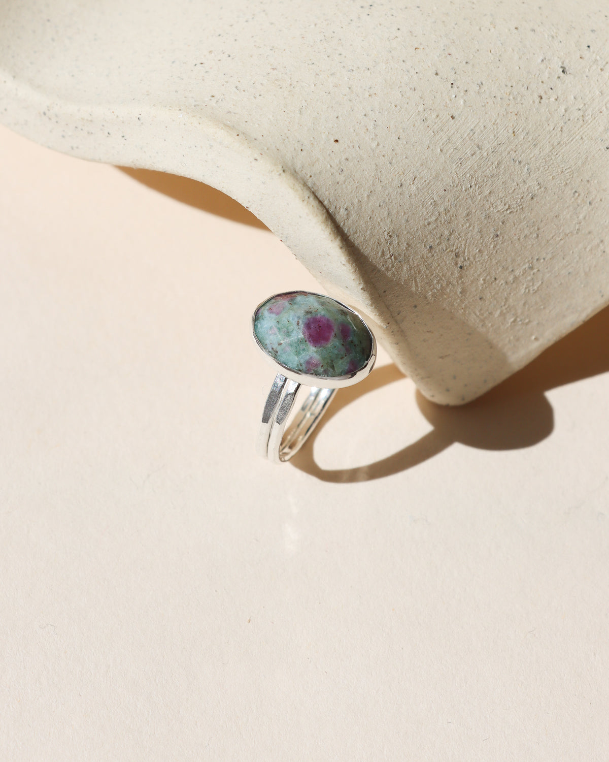 Oval green stone ring with pink and purple accents set in silver, photographed in warm sunlight against a cream background beside a textured ceramic form.
