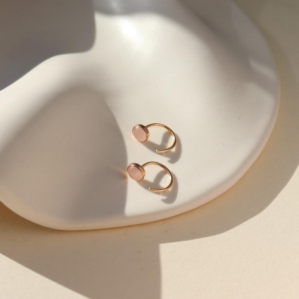 Our rose quartz Huggies feature a 5mm rose quartz gemstone. These earrings are made to hug your ear featuring a curved 14k gold fill post that will wrap around your ear. The earrings are on a white tray. 