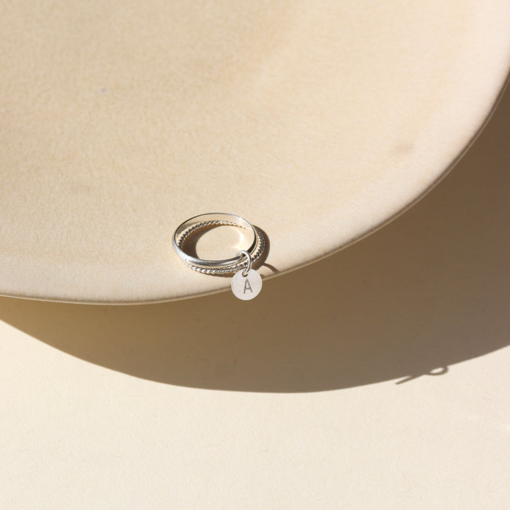 A smooth band stacked with a spiral sterling silver band, with a small "A" stamped disc attached to both rings. 