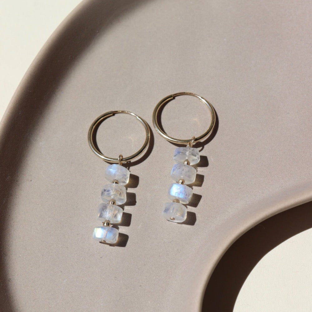 14k gold fill Selene Hoops laid on a tan paper in the sunlight. These Earrings feature our 16mm Goldie hoops along with 4 Moonstone Faceted gemstones stacked onto of each other