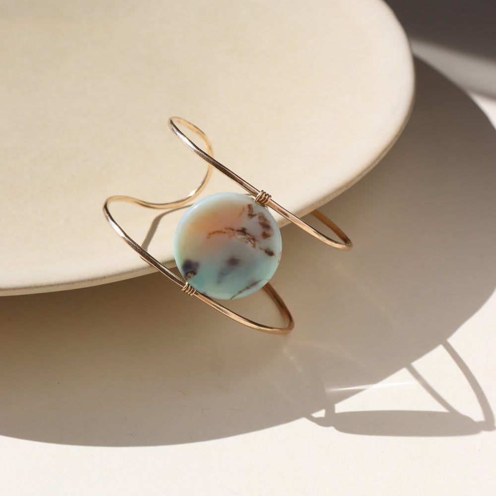 The siren cuff features a Terra Agate Gemstone wire wrapped to a 14k gold fill wide cuff. The cuff is displayed against a cream plate in the sunlight. 